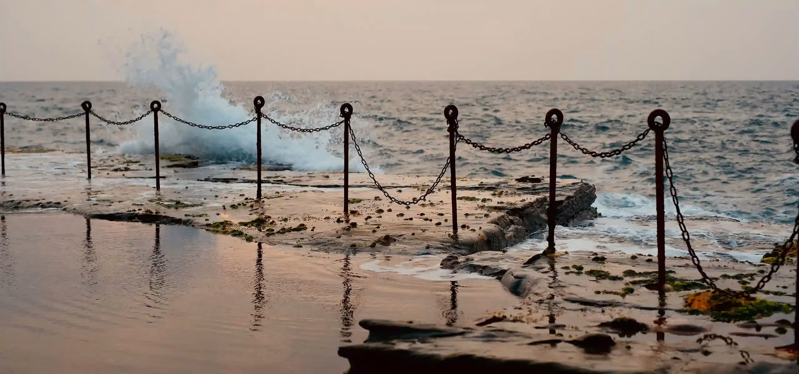 A wave breaking over the edge of an ocean rock pool, its chain railing on iron posts, at dusk
