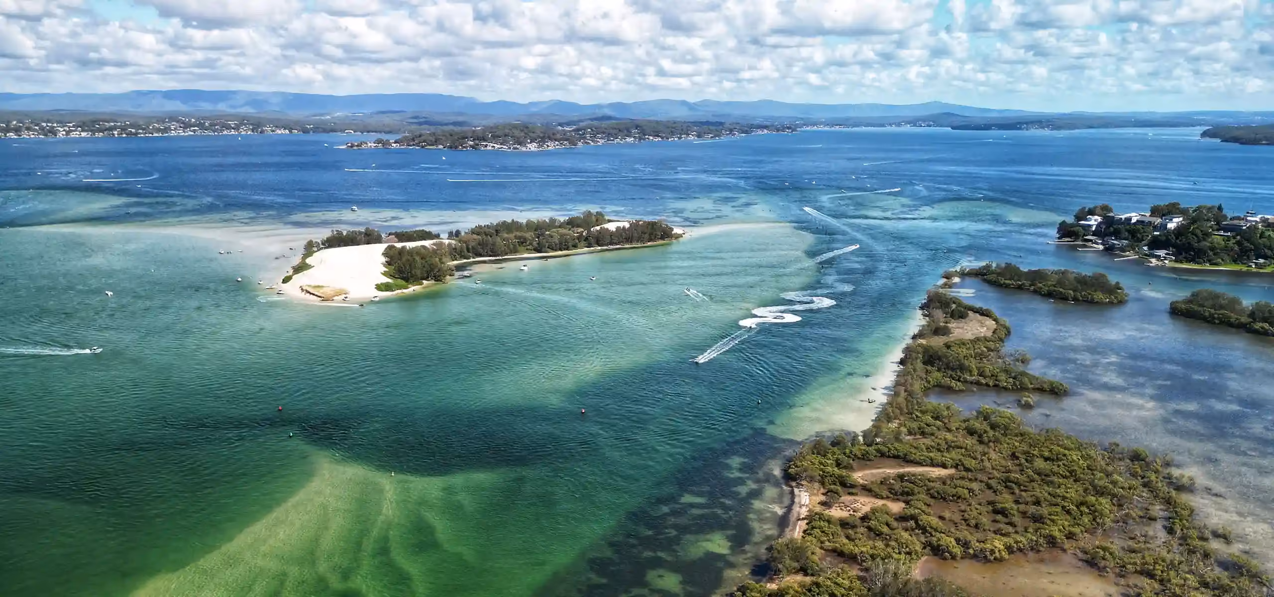 An aerial view of a shallow lake channel with a sandy wooded island, boats cutting wakes, and a long range of hills behind